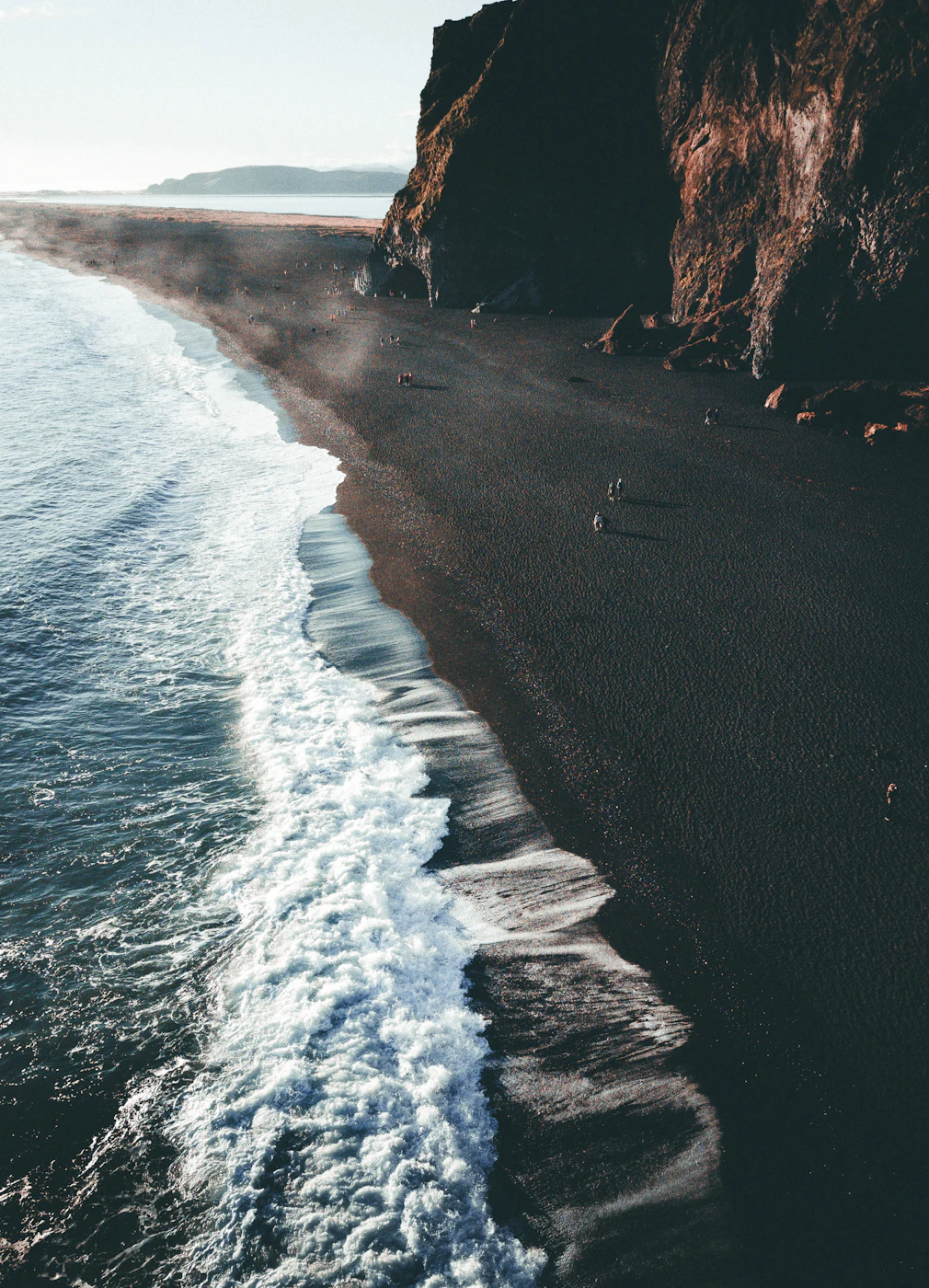 Surf rolling onto a long black beach below a cliff